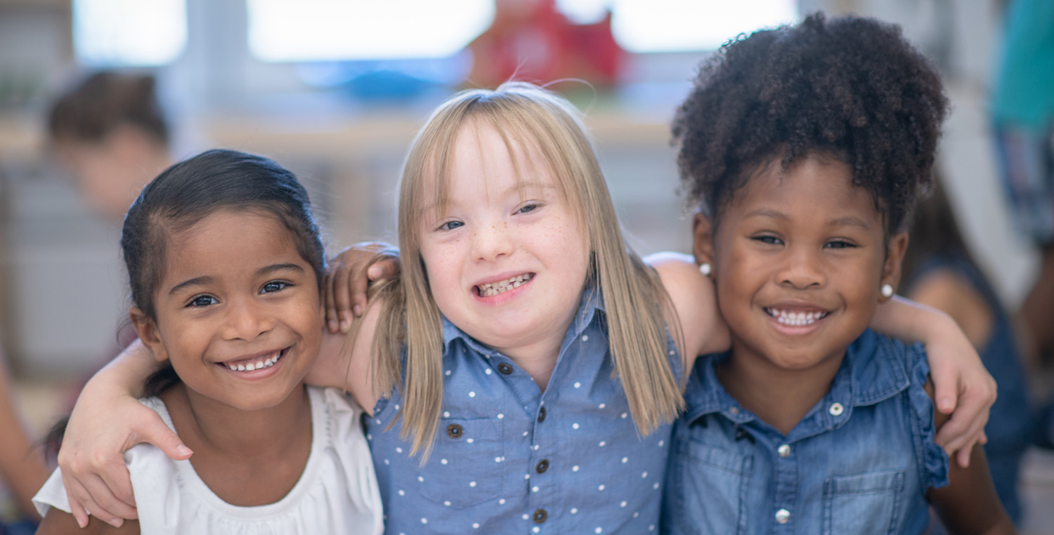 Three multi ethnic preschool girls in their classroom. One of the girls (in the middle) is of Caucasian ethnicity and has Down syndrome. The other girls are of African and mixed race ethnicity.