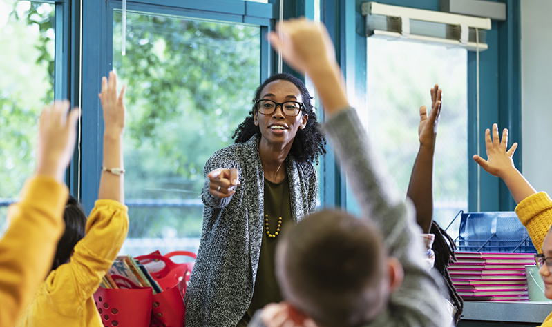 A multiracial group of elementary students in class raising their hands to answer a question. The focus is on their teacher, an African-American and Hispanic woman in her 30s, who is standing in front, facing them, smiling and pointing to a student. Most of the students are 9 years old.